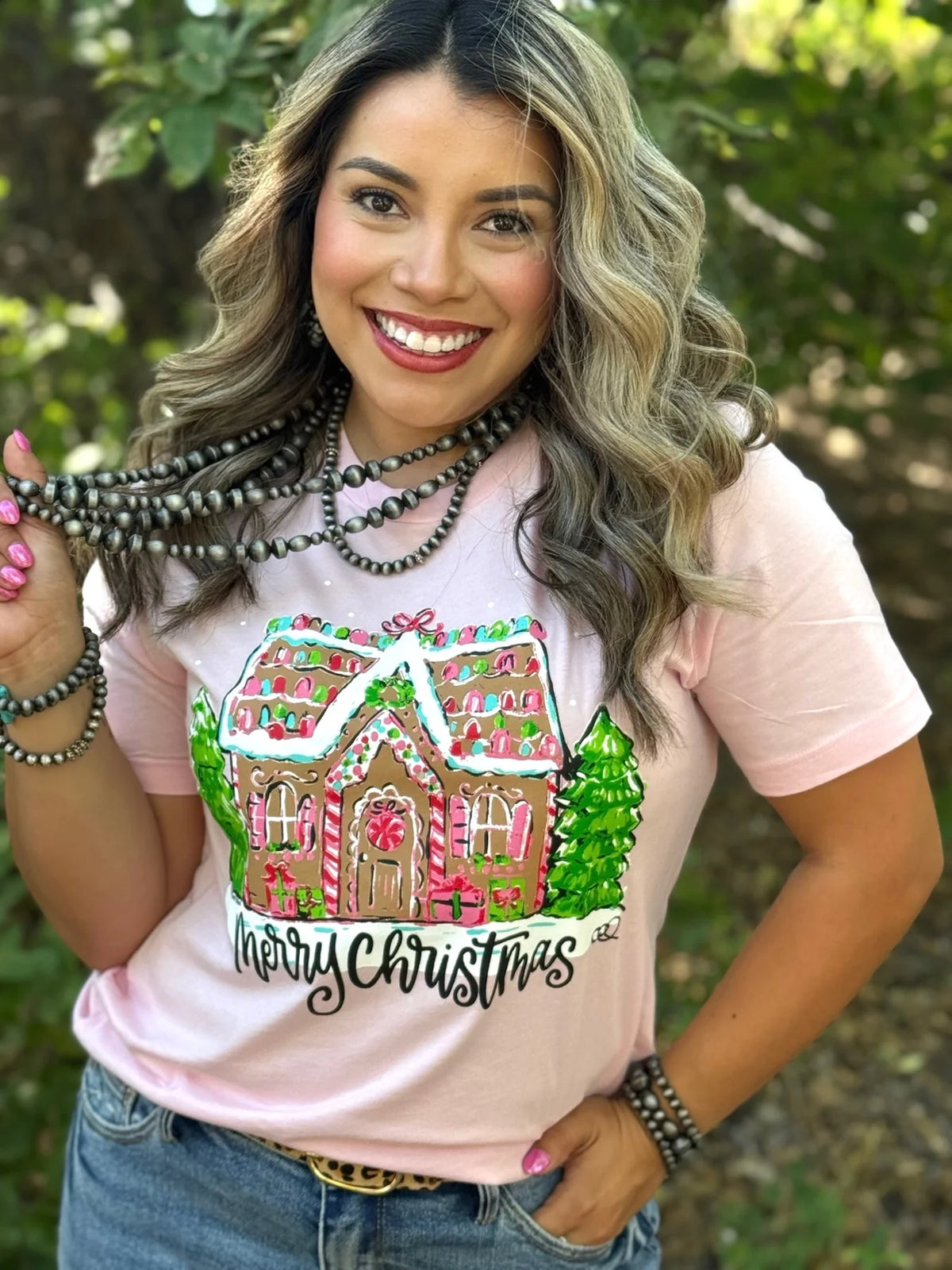Woman wearing a pink t-shirt with a gingerbread house design and 'Merry Christmas' text, standing outdoors.